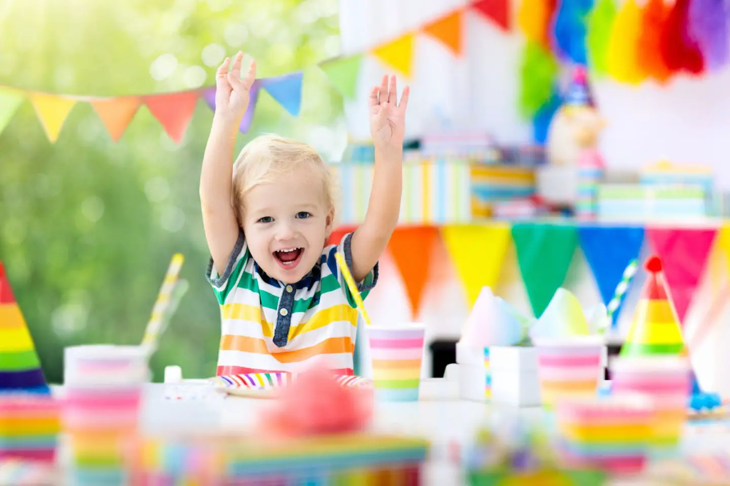 Kids birthday party. Child blowing out candles on colorful cake. Decorated home with rainbow flag banners, balloons. Farm animals theme celebration. Little boy celebrating birthday. Party food.